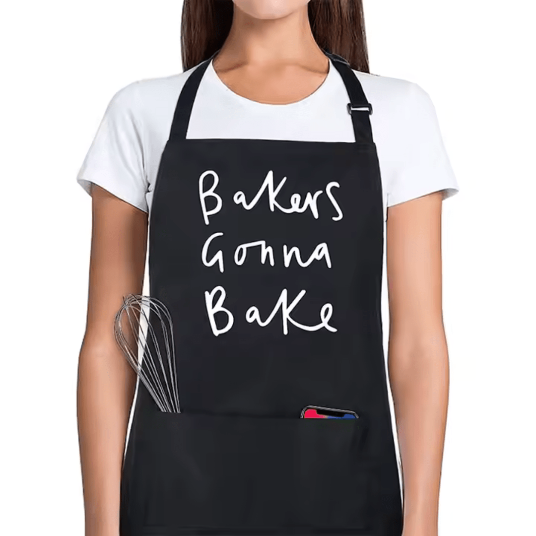 Black cotton apron with white text reading ‘Bakers Gonna Bake’ worn by woman in kitchen.