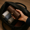 Brown leather watch roll case placed in black duffel bag with rolled shirts and sunglasses, luxury travel style.