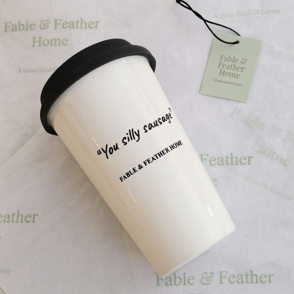 White ceramic travel mug with black lid, featuring the phrase “You silly sausage” and the Fable & Feather Home logo, displayed on branded tissue paper.
